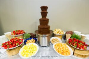 4 tier dark chocolate fountain with dipping items around - pineapple, strawberries, and madeleines