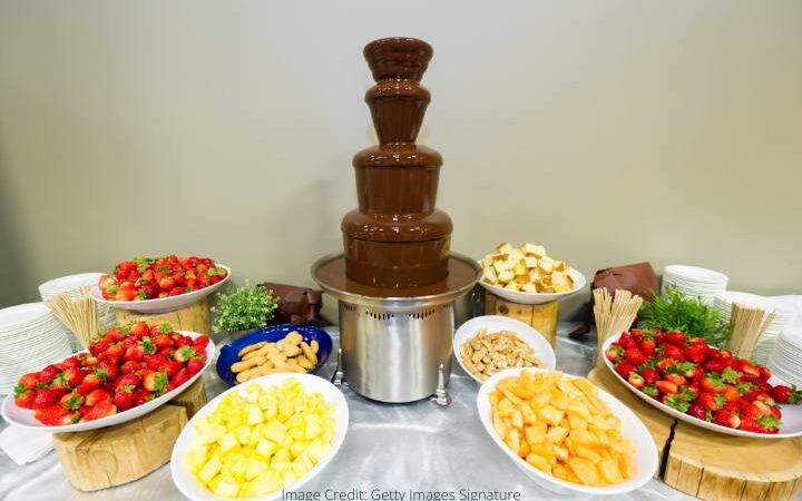 4 tier dark chocolate fountain with dipping items around - pineapple, strawberries, and madeleines