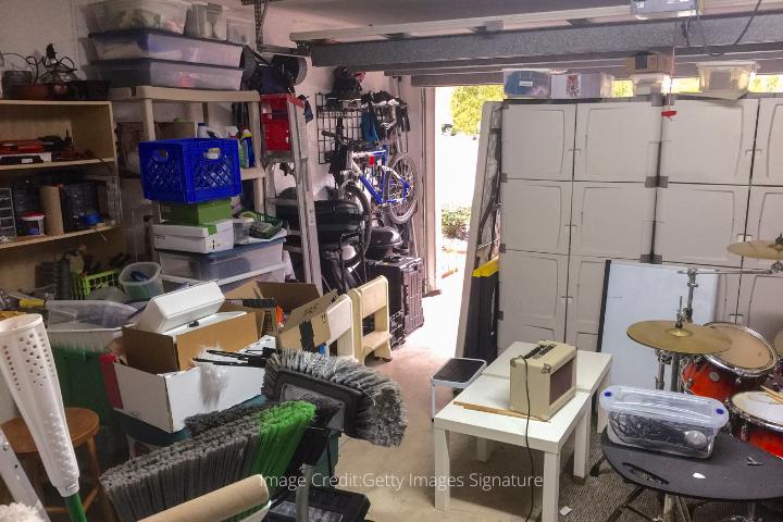 messy garage interior with brooms, a beige amp on a white table, a red drumset in front of beige containers that are stacked up 5 feet high, shelves of cables, plastic containers, and shoe boxes, a bike on a rack