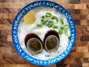 image: bowl of black, preserved duck egg cut in half over rice porridge, slice of ginger, and sliced green onions in a blue bowl on a wood cutting board