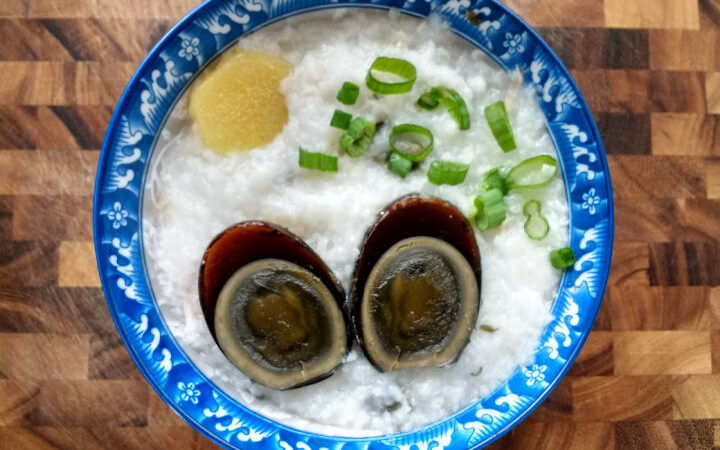 image: bowl of black, preserved duck egg cut in half over rice porridge, slice of ginger, and sliced green onions in a blue bowl on a wood cutting board