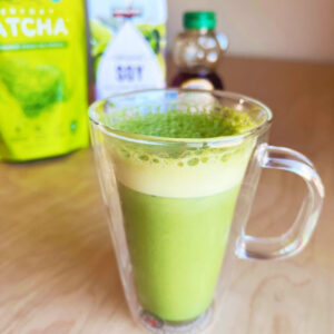 glass of iced matcha latte with ingredients in background - matcha powder, soy milk, and honey